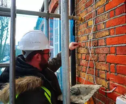 Worker inspecting brick wall during restoration, wearing hard hat and safety gear, with scaffolding and tools visible, highlighting building maintenance and repair challenges.