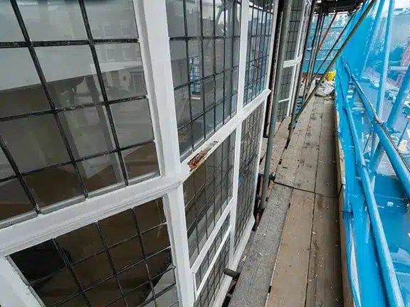 Sash window restoration in progress, showcasing detailed wooden frames and grid patterns, with scaffolding and protective covering in a period home setting.