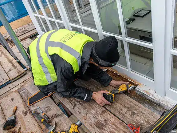 Worker repairing timber windows on a scaffold, wearing high-visibility jacket, surrounded by tools, demonstrating restoration techniques for period properties.