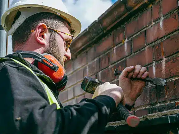 Worker repairing masonry with chiselling tools and safety gear, focused on repointing brickwork for heritage restoration.