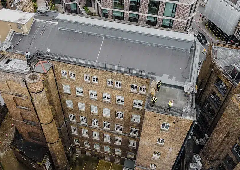 Aerial view of a large building with maintenance workers in high-visibility jackets inspecting the roof, showcasing the efficiency of drone roof surveys by Environ Restoration Services.