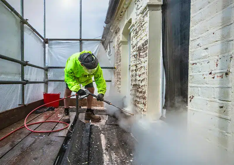 Worker in high-visibility jacket using pressure washer to clean historic building facade, surrounded by scaffolding and protective sheeting, highlighting period property conservation efforts in London.