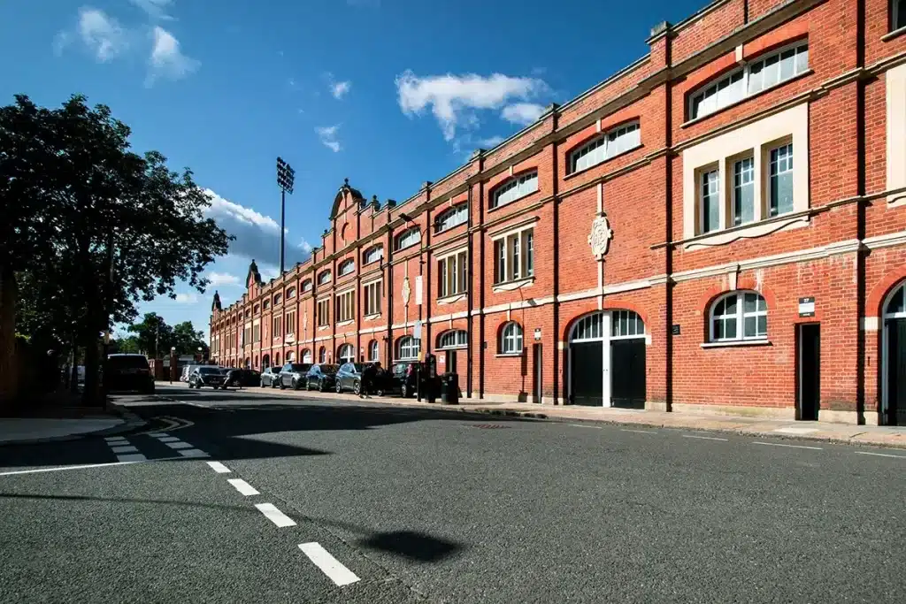 Historic red brick building along a London street, featuring large windows, decorative architectural elements, and parked cars, with a clear blue sky above.