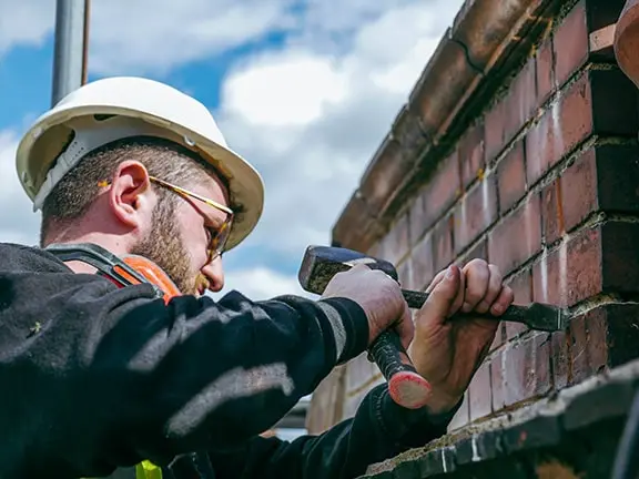 Worker repairing a stone wall with a hammer and chisel, wearing a hard hat and safety glasses, against a backdrop of blue sky and clouds, illustrating expert stone wall restoration techniques.