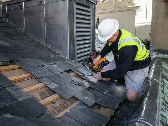 Worker repairing a slate roof on a period building, wearing a hard hat and high-visibility vest, demonstrating expert craftsmanship in restoration services.