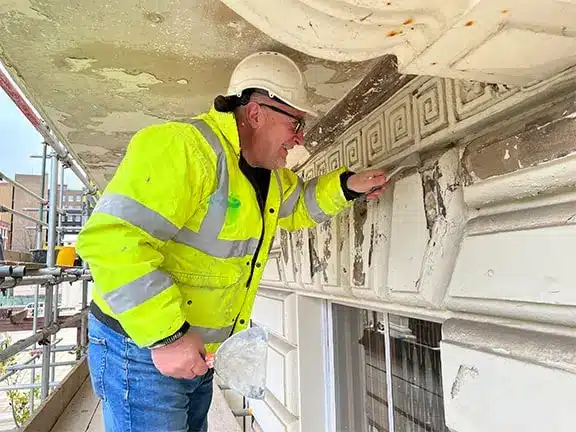 Worker in high-visibility jacket restoring decorative architectural details on a listed building, showcasing expertise in historical restoration techniques.