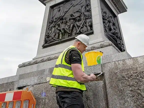 Worker in safety gear restoring a listed building, applying finishing touches to a stone structure with intricate carvings, emphasizing quality restoration techniques.