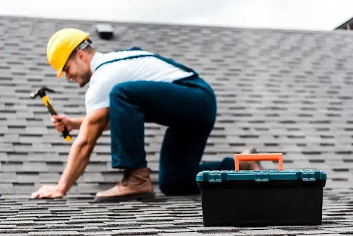 Roofer in yellow hard hat and blue overalls repairing a grey shingle roof, with a toolbox nearby, illustrating expert roofing services for period property restoration in London.