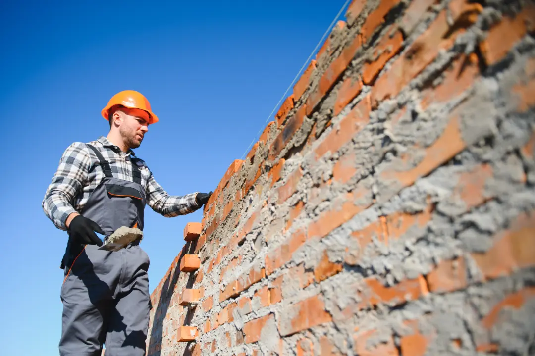 Construction worker in hard hat applying mortar to brick wall, illustrating professional repointing techniques relevant to optimal seasons for brickwork restoration.
