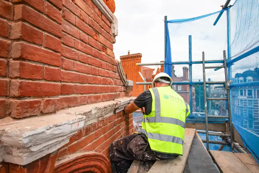 Construction worker in high-visibility vest repointing brickwork on a London building, with scaffolding and cityscape in the background, illustrating property maintenance and restoration services.
