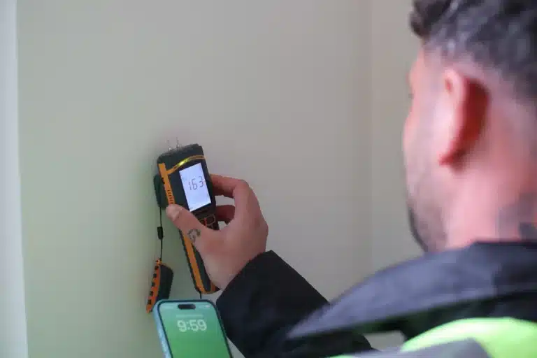 Technician using a damp meter to assess moisture levels on a wall, with a smartphone displaying the time, highlighting professional damp diagnosis services in London.