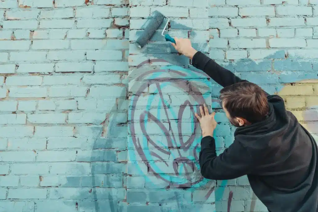 Person using a paint roller to cover unwanted graffiti on a blue brick wall, illustrating graffiti removal techniques for masonry surfaces.