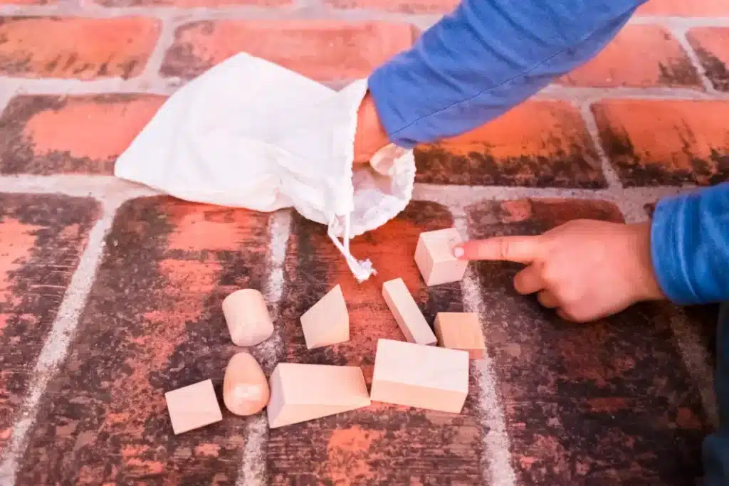 Child's hand pointing at wooden building blocks scattered on a brick surface, with a white bag partially visible.