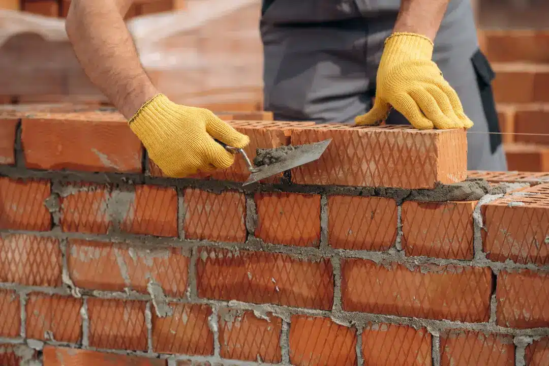Person in yellow gloves laying bricks with trowel, demonstrating brickwork techniques for cleaning and restoration.