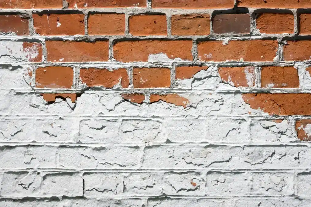 Brick wall with peeling white paint revealing underlying red bricks, illustrating paint removal challenges and potential damage to brickwork.